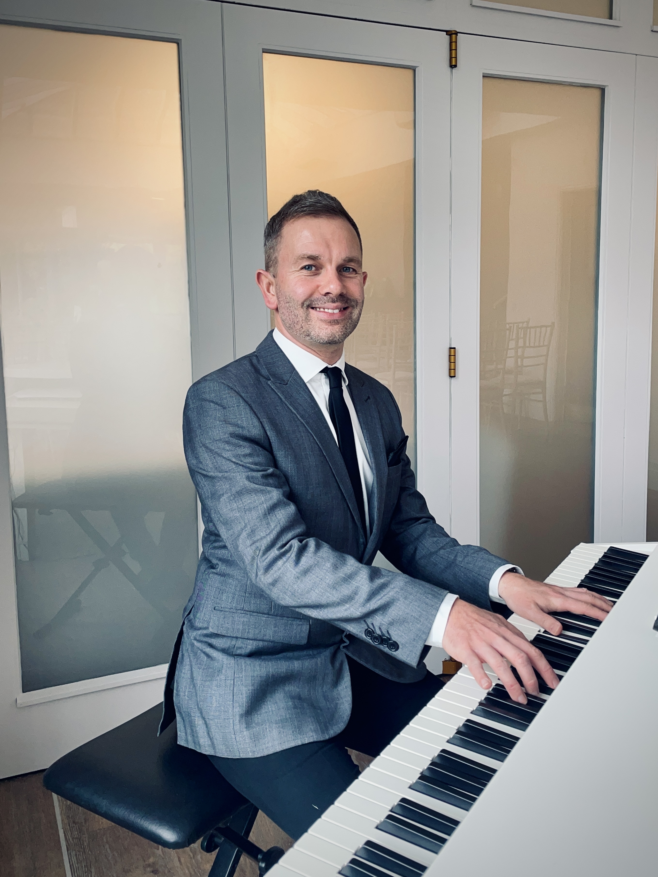 The Lake District Pianist smiles while playing a white baby grand piano for a wedding ceremony at Broadoaks Country House Hotel. He wears a charcoal jacket over a white shirt, black tie and black trousers.
